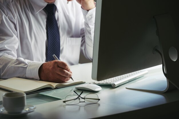 Mid sectie van onherkenbare man in formalwear maken van aantekeningen op werkplek computer