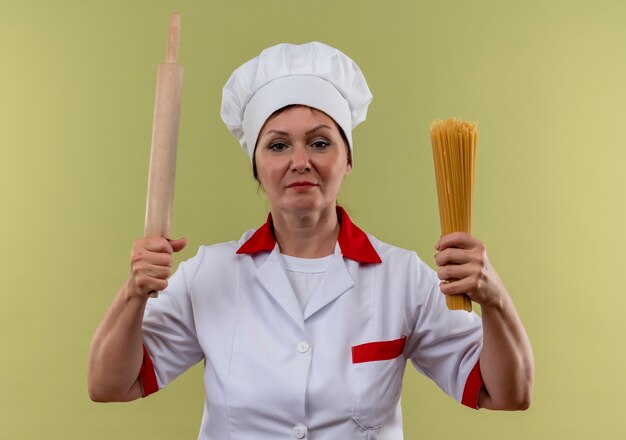 middelbare leeftijd vrouwelijke kok in chef-kok uniform met deegroller en spaghetti op geïsoleerde groene muur