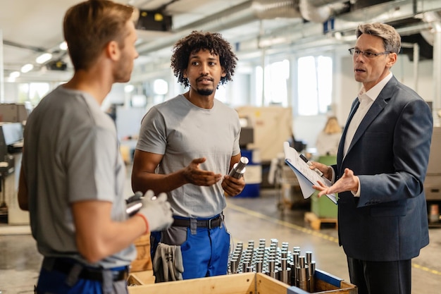 Middenvolwassen manager in gesprek met metaalbewerkers tijdens het uitvoeren van kwaliteitscontrole van gefabriceerde staafcilinders in een fabriek