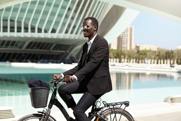 Milieuvriendelijke zwarte manager in formeel pak en zonnebril die vooruit kijken terwijl ze met de fiets naar het werk gaan in een stedelijke omgeving, gelukkig lachend. Zaken, lifestyle, transport en mensen