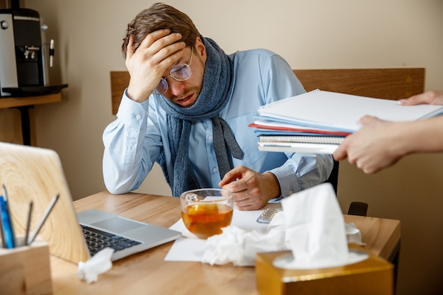 Misselijk en moe voelen. Gefrustreerd triest ongelukkig zieke jongeman zijn hoofd masseren zittend op zijn werkplek op kantoor. De seizoensgriep, pandemische influenza, ziektepreventie concept