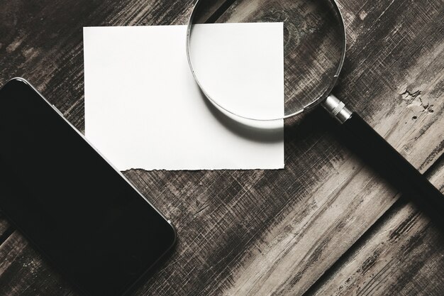 Mobiele smartphone, vergrootglas en vel wit papier geïsoleerd op zwarte boerderij houten tafel Mysterieus detective spelconcept. Zijaanzicht van de close-up