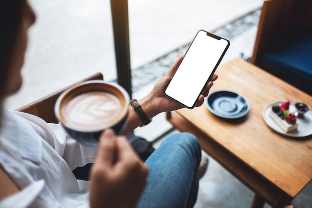 Mockup-afbeelding van een vrouw die een mobiele telefoon vasthoudt met een leeg wit desktopscherm terwijl ze koffie drinkt in café Premium Foto
