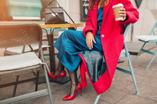 Mode-accessoires van stijlvolle vrouw zitten in stad straat café in rode jas koffie drinken dragen blauwe zijden jurk, schoenen met hoge hakken