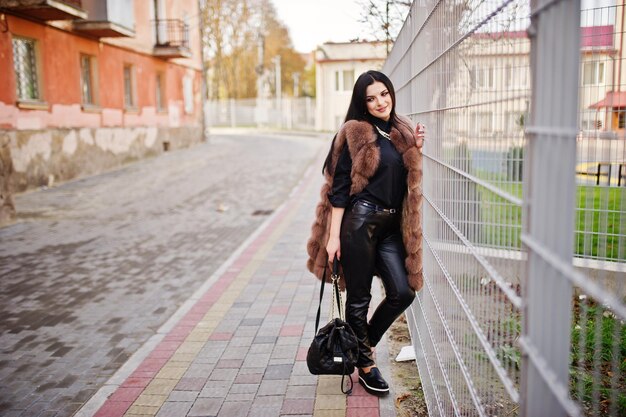 Mode-buitenfoto van prachtige sensuele vrouw met donker haar in elegante kleding en luxe mouwloze bontjas tegen ijzeren hek in herfststad