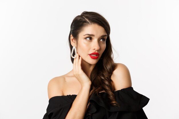 Mode en beauty concept. Close-up van elegante vrouw in zwarte jurk, oorbellen tonen en op zoek naar sensuele, rode lippenstift en make-up op, witte achtergrond