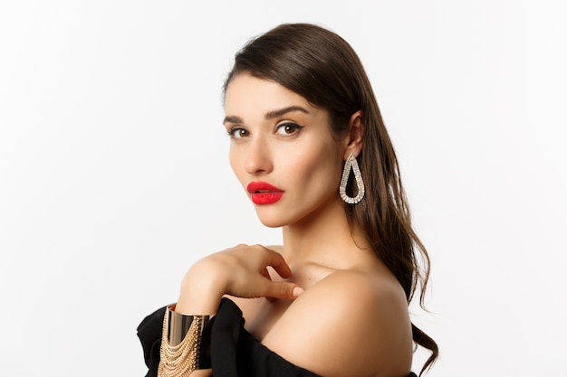 Mode en beauty concept. Close-up van elegante vrouw met rode lippen, make-up en oorbellen, camera kijken zelfverzekerd, staande op witte achtergrond.