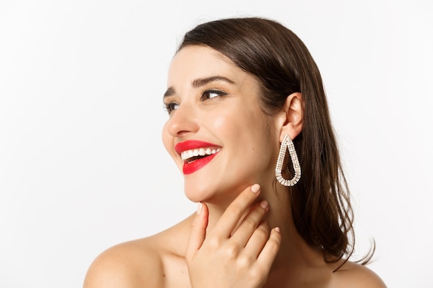 Mode en beauty concept. Headshot van prachtige brunette vrouw met rode lippenstift, oorbellen, lachen en naar links kijken, staande op een witte achtergrond.