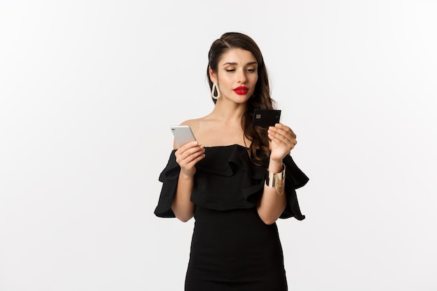 Mode- en winkelconcept. vrouw in make-up en zwarte jurk online bestellen, betalen met creditcard en mobiele telefoon, witte achtergrond.