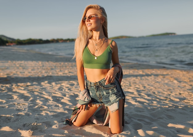 Mode foto van sexy blonde vrouw in groene crop top en jeans poseren op tropisch strand.