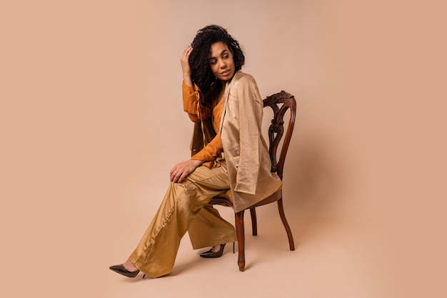 Mode portret van aantrekkelijke vrouw met bruine huid met perfect krullende haren in elegante oranje blouse en zijden broek zittend op vintage stoel op beige muur.