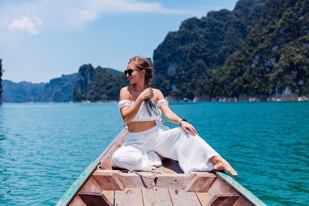 Mode portret van een jonge vrouw in witte top en broek op vakantie, op Thaise houten zeilboot. Reis concept. Vrouwtje in Khao Sok National park.