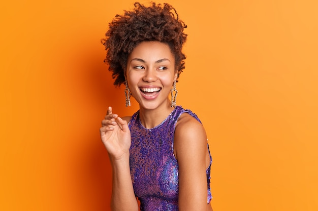 Mode portret van gelukkige stijlvolle vrouw in lichte kleren oorbellen glimlacht in grote lijnen kijkt opzij geïsoleerd over oranje muur