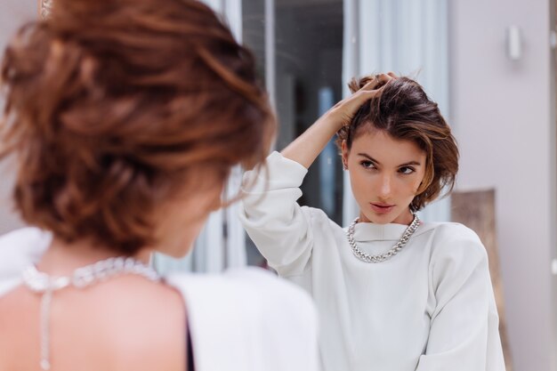 Mode portret van jonge blanke vrouw professioneel model in witte blazer en zilveren ketting kijken in spiegel luxevilla