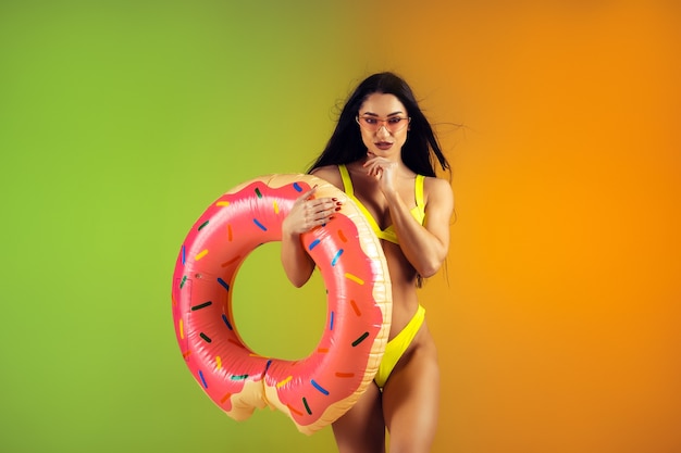 Mode portret van jonge, fitte en sportieve vrouw met rubberen donut in stijlvolle gele badmode op het perfecte lichaam van de gradiëntmuur, klaar voor de zomer summer