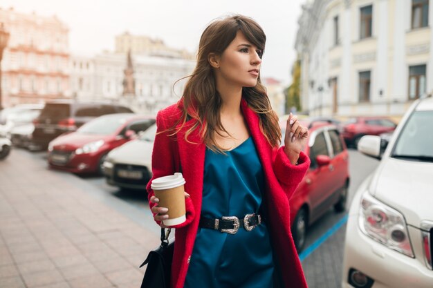 Mode portret van jonge mooie stijlvolle vrouw wandelen in de stad straat in rode jas, herfst stijl trend, koffie drinken, glimlachen, gelukkig, blauwe zijden jurk dragen