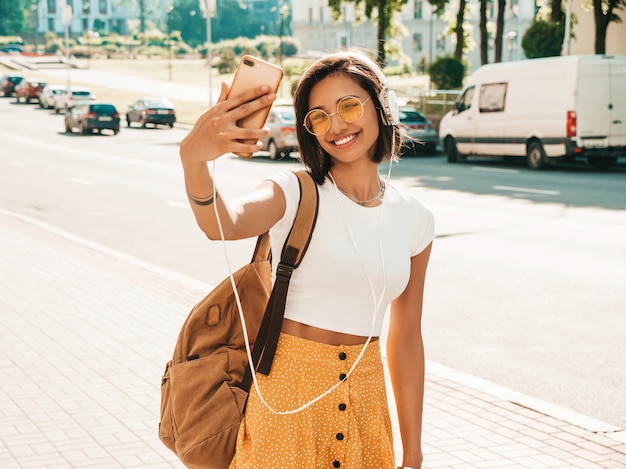 Mode portret van jonge stijlvolle hipster vrouw lopen in de straat. Meisje selfie maken. Glimlachend model geniet van haar weekenden met rugzak. Vrouw luisteren naar muziek via de koptelefoon