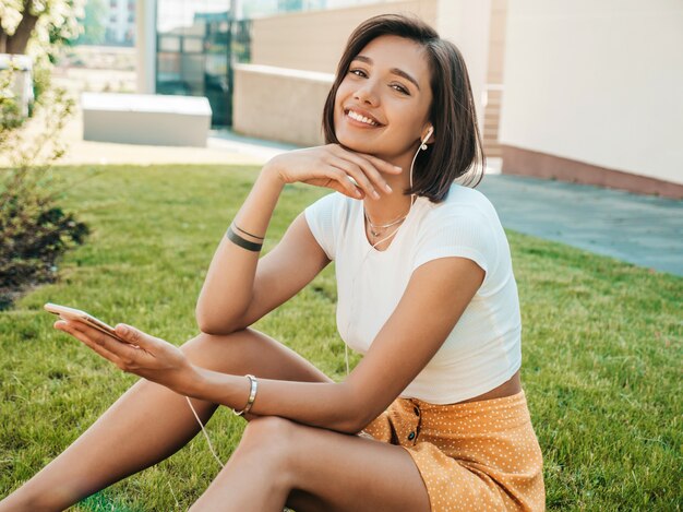 Mode portret van jonge stijlvolle hipster vrouw. Meisje draagt schattige trendy outfit. Glimlachend model geniet van haar weekenden, zittend in het park. Vrouw luisteren naar muziek via de koptelefoon