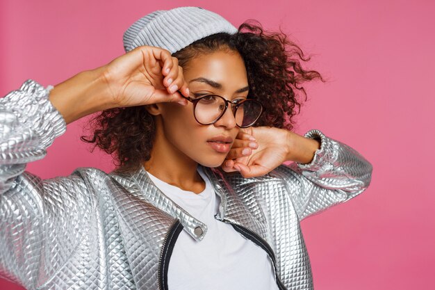 Mode portret van mix race vrouw met bruine huid en krullend Afrikaans kapsel op levendige roze achtergrond. Het dragen van zilveren winterjas en grijze hoed.