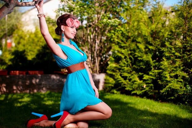 mode portret van mooie jonge vrouwelijke model dame vrouw met kapsel in heldere blauwe jurk poseren buitenshuis zitten in groen gras in de buurt van struik met bloemen in haar