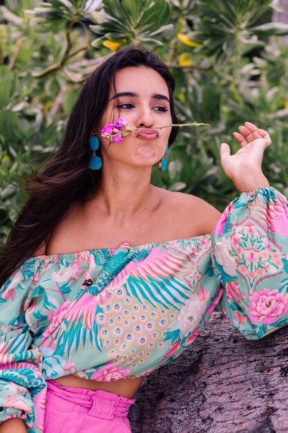 Mode portret van stijlvolle vrouw in kleurrijke print top met lange mouwen en roze broek op strand, tropische achtergrond.