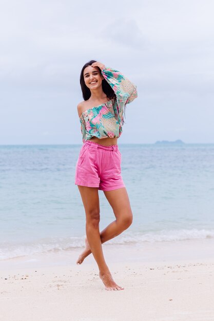 Mode portret van stijlvolle vrouw in kleurrijke print top met lange mouwen en roze broek op strand, tropische achtergrond.