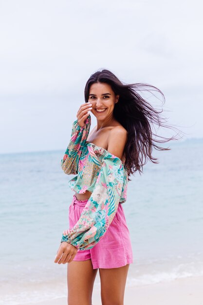 Mode portret van stijlvolle vrouw in kleurrijke print top met lange mouwen en roze broek op strand, tropische achtergrond.