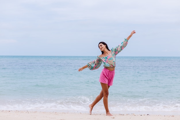 Mode portret van stijlvolle vrouw in kleurrijke print top met lange mouwen en roze broek op strand, tropische achtergrond.