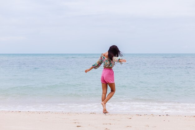 Mode portret van stijlvolle vrouw in kleurrijke print top met lange mouwen en roze broek op strand, tropische achtergrond.