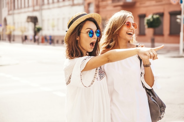 Mode portret van twee jonge stijlvolle hippie brunette en blonde vrouwen modellen in zonnige zomerdag in witte hipster kleding poseren. wijzend op winkelverkopen