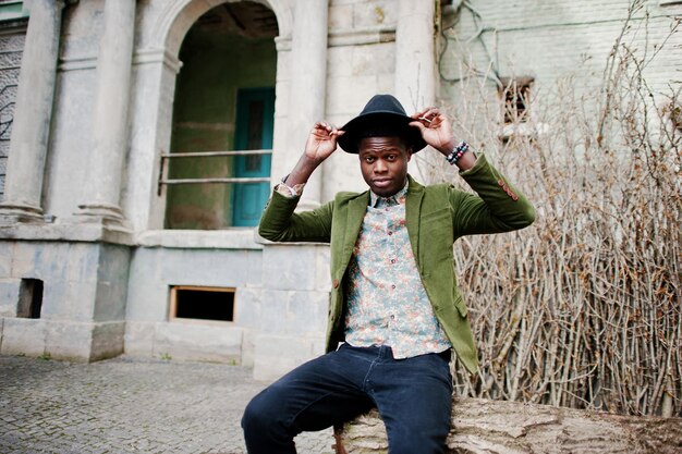 Mode portret van zwarte Afro-Amerikaanse man op groen fluwelen jasje en zwarte hoed zittend op gekapte boom achtergrond oude vintage huis met boog en kolommen