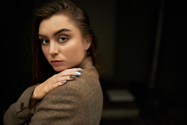 Mode, stijl en beauty concept. Profiel shot van ernstige zelfverzekerde succesvolle jonge Europese vrouw met bruin haar en gepolijste nagels stijlvolle jas dragen, hand houden op haar schouder