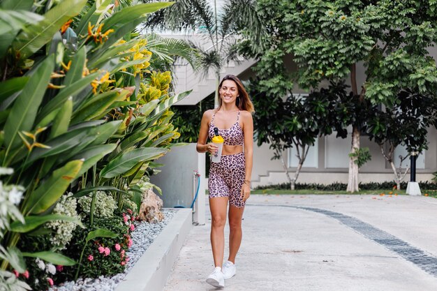 Mode stijlvolle jonge Kaukasische fit sportieve vrouw in luipaard cami top en biker shorts buiten houdt eiwit shaker, fles water, in de hand