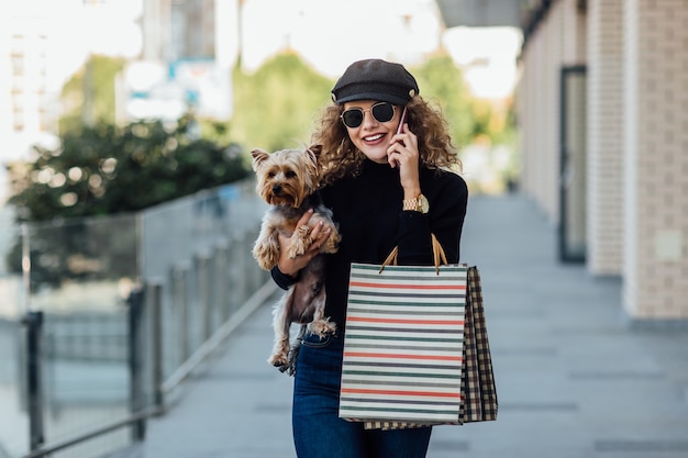 Mode wandeling vrouw met lang krullend haar houdt kleine hond en boodschappentassen Mooi meisje knuffels hondje Glimlachende aantrekkelijke vrouw met Yorkshire terrier Meisje met hond in handen en verkoop concept