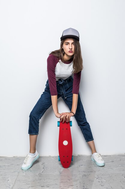 Modern schaatsermeisje die rood skateboard op de vloer houden die op witte muur wordt geïsoleerd