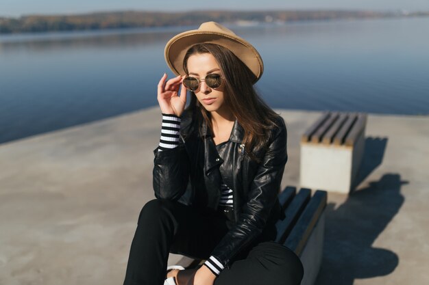 Moderne jonge model meisjesvrouw zitten op een bankje in de herfstdag aan de waterkant van het meer