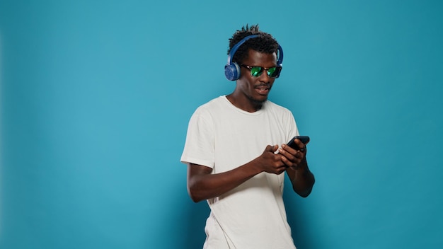 Moderne persoon met koptelefoon die naar muziek luistert en smartphone gebruikt met touchscreen. Vrolijke man met zonnebril dansen, genieten van lied op headset terwijl hij mobiele telefoon en kopje koffie vasthoudt.