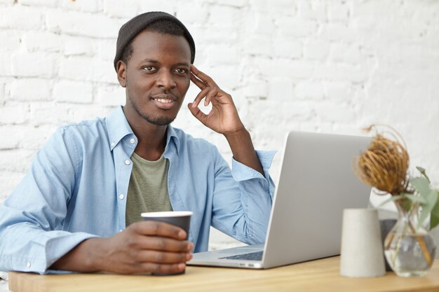 Moderne stedelijke levensstijl en technologieën concept. Aantrekkelijke jonge Afro-Amerikaanse mannelijke freelancer met hoed met koffiepauze tijdens het werken op afstand op laptop pc, op zoek nadenkend of dromerig