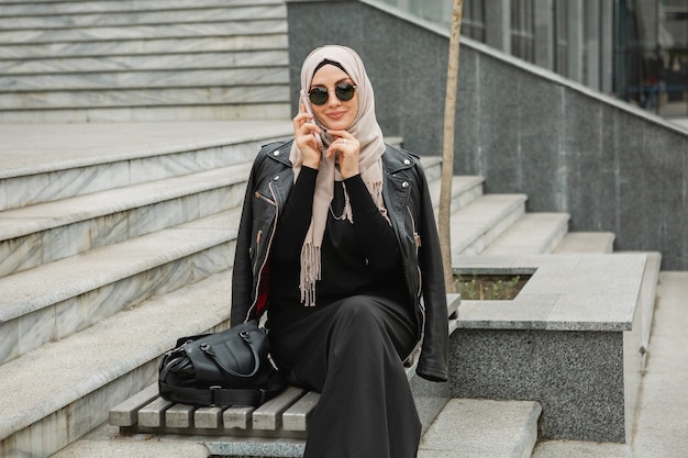 Moderne stijlvolle moslimvrouw in hijab, leren jas en zwarte abaya die in de stad loopt en op smartphone praat