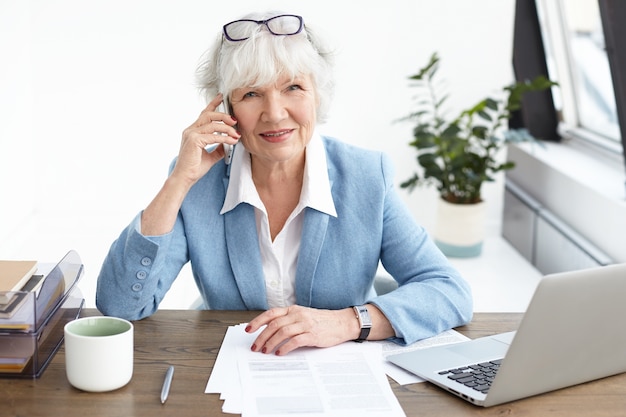 Moderne technologie, communicatie en baanconcept. Hoge hoekmening van succesvolle ervaren oudere zakenvrouw dragen pak en stijlvolle accessoires met telefoongesprek met behulp van mobiel