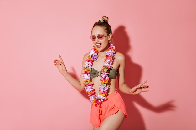 Moderne vrouw in stijlvolle zonnebril, cool badpak roze korte broek en ketting van bloemen glimlachend op geïsoleerde achtergrond