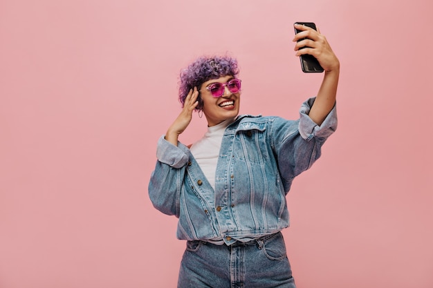 Moderne vrouw met paars haar in coole zonnebril maakt foto op roze. Glimlachende dame in brede lichte kleren neemt selfie.