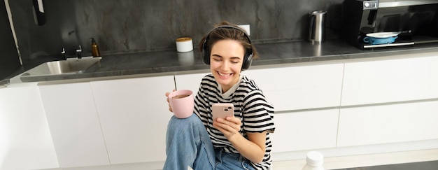 Moderne vrouwelijk model glimlacht met behulp van mobiele telefoon thee drinken en ontbijt eten in de keuken