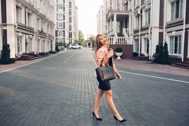 Modieus blond meisje met lang haar, wandelen in zwarte jurk rond Britse wijk. Ze houdt koffie vast