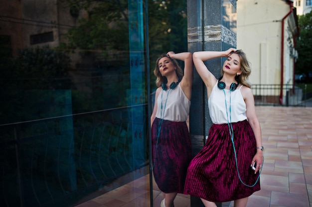 Modieus en mooi blond model meisje in stijlvolle rode fluwelen velours rok witte blouse poseerde met telefoon en koptelefoon tegen ramen van stadsgebouw