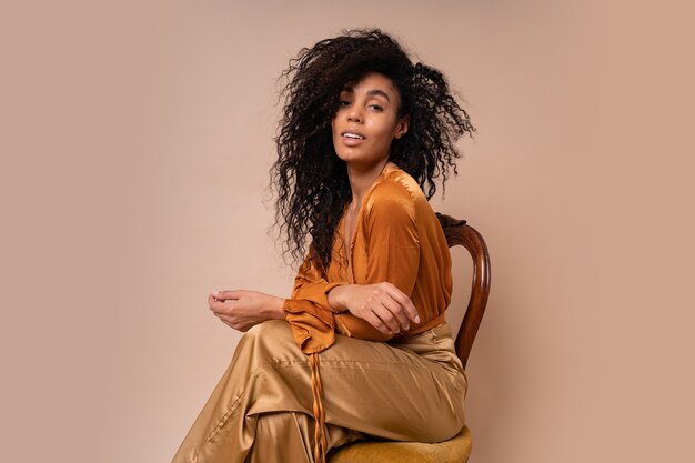 Modieus jong verleidelijk Afrikaans model met perfect krullende haren in elegante oranje blouse en zijden broek zittend op vintage stoel beige muur.