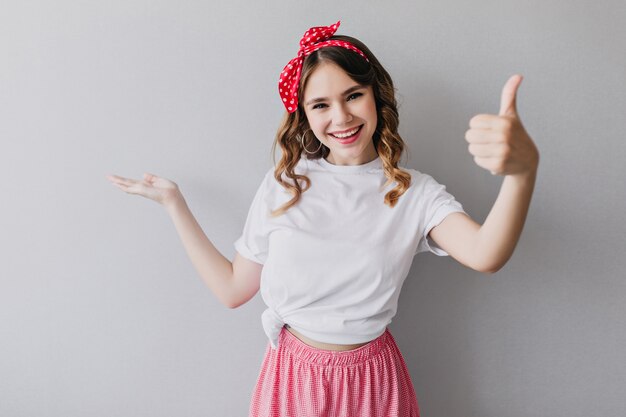 Modieus meisje dat met rood lint met omhoog duim glimlacht. portret van fascinerende dame in trendy zomerkleren.