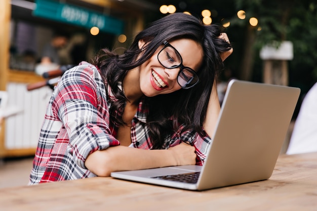 Gratis foto modieus meisje in glazen die positieve emoties uitdrukken tijdens het werken met laptop. outdoor portret van schattige brunette student huiswerk.