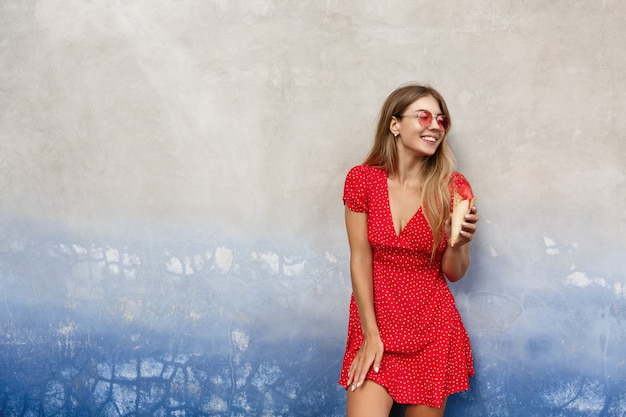 Modieus stedelijk meisje in rode zonnebril en zomerjurk, buitenshuis eten van ijs, leunend op een betonnen muur en wegkijken