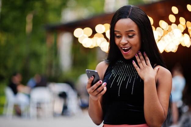 Modieuze Afro-Amerikaanse vrouw in perzik broek en zwarte blouse kijken naar mobiele telefoon